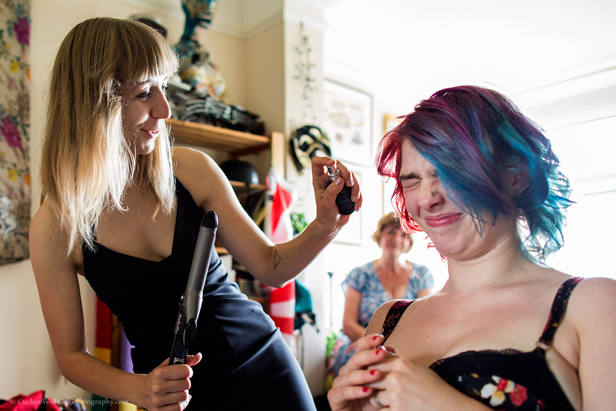 An alternative bride with red and blue hair squints as a bridesmaid sprays here face from a bottle.