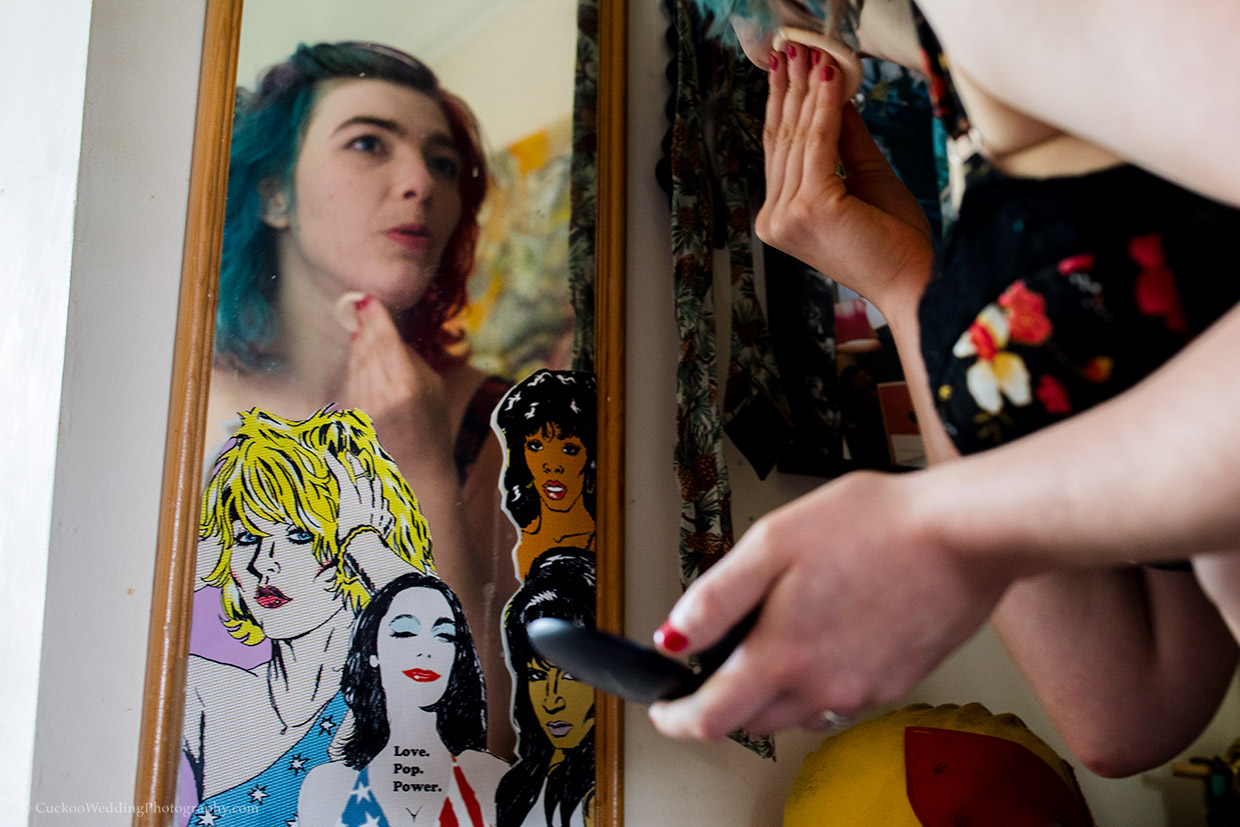 An quirky bride with blue hair is applying make up in a mirror with pictures of confident women on it.