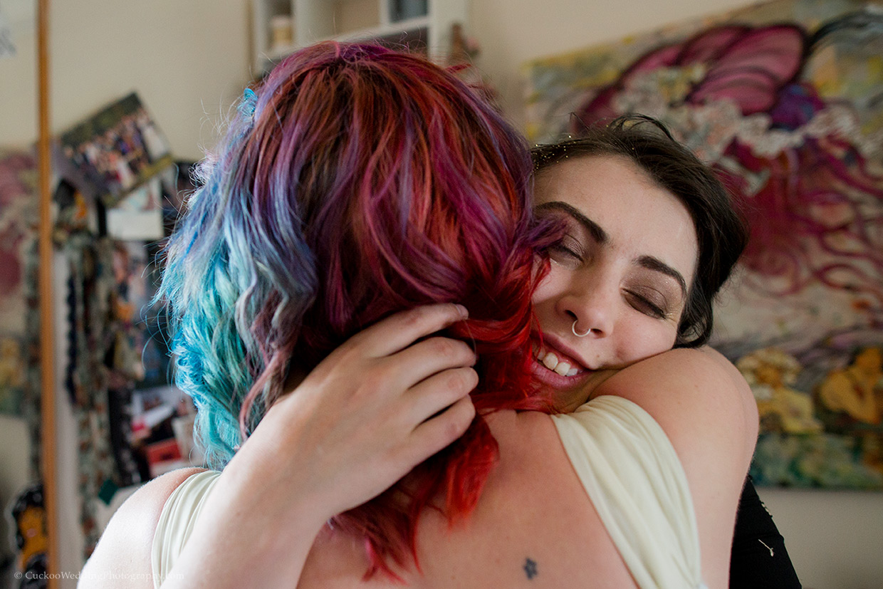 A bridesmaid hugs a quirky bride with red and blue dyed hair. She has her eyes closed and is very happy.