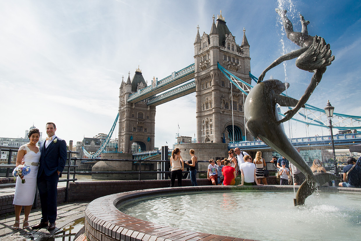 Unique bride and groom are smiling in font of Tower Bridge in London, there is a fountain with a dolphin in it, it's a sunny day.
